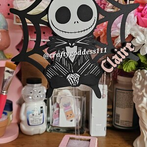 May include: A decorative birthday cake topper featuring Jack Skellington from The Nightmare Before Christmas. The black and white design includes the text "Happy Birthday" and the name "Claire". The topper is on a pink and white base.