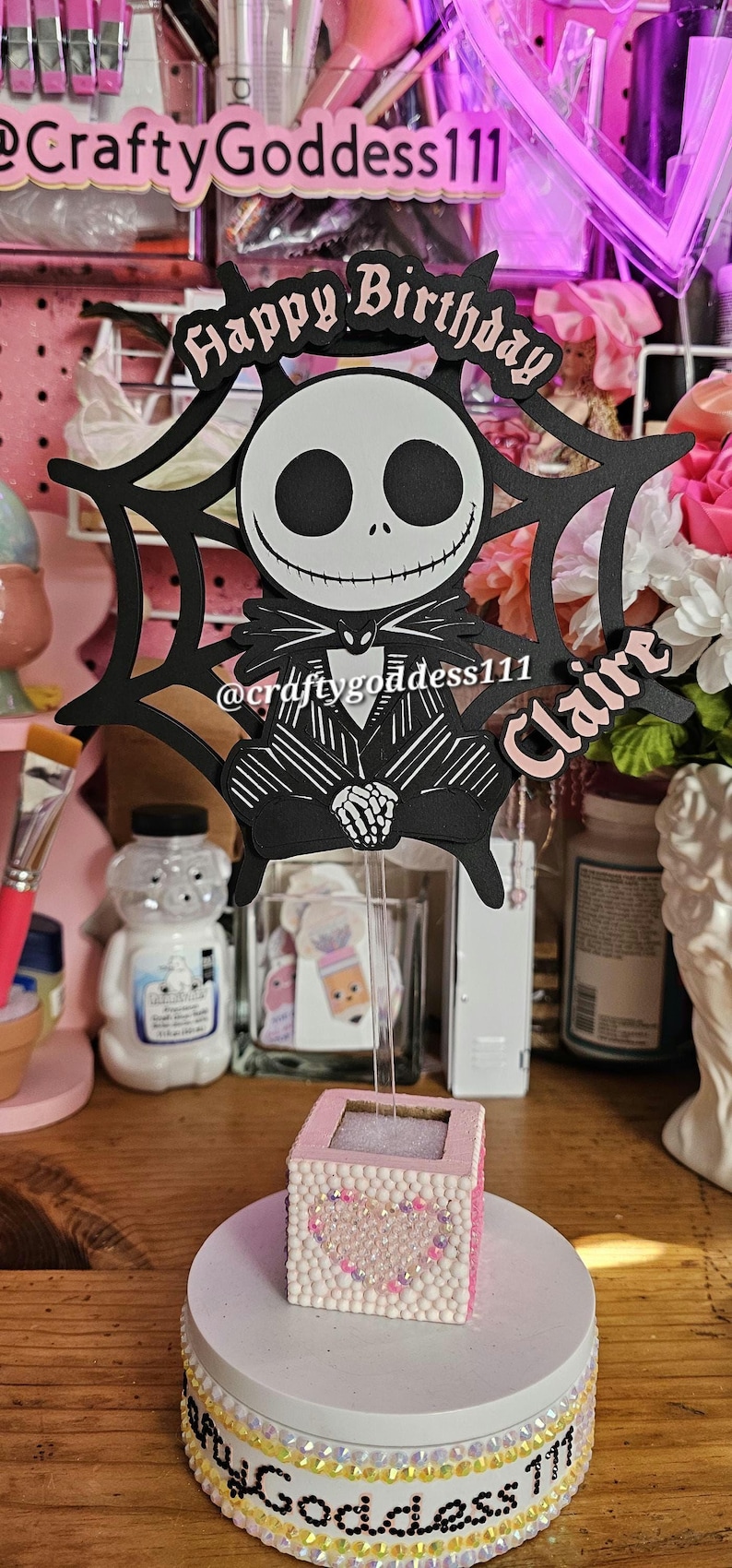 May include: A decorative birthday cake topper featuring Jack Skellington from The Nightmare Before Christmas. The black and white design includes the text "Happy Birthday" and the name "Claire". The topper is on a pink and white base.