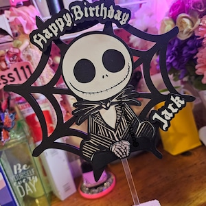 May include: A black and white Jack Skellington cake topper with the words "Happy Birthday Jack". The topper features a spiderweb design and is attached to a clear plastic stick for insertion into a cake.