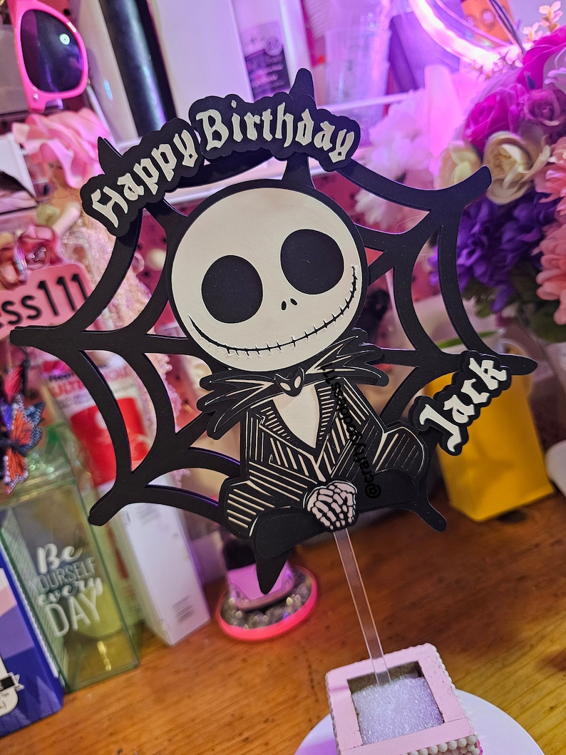 May include: A black and white Jack Skellington cake topper with the words "Happy Birthday Jack". The topper features a spiderweb design and is attached to a clear plastic stick for insertion into a cake.
