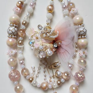 May include: A whimsical beaded keychain featuring multiple strands of iridescent and pearl-like beads in shades of pink, cream, and gold. The keychain includes a fairy charm with a pink wing and gold accents, and is attached to gold-colored clasps.