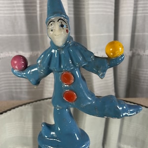May include: A blue and white ceramic clown candle with a red nose and two orange buttons. The clown is holding a yellow ball in one hand and a pink ball in the other.