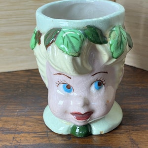 Puede incluir: Una taza de cerámica con forma de cara de mujer con hojas verdes en la cabeza. La taza tiene un fondo blanco con detalles rosas y verdes. Los ojos de la mujer son azules y sus labios son rojos.
