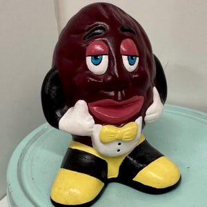 Puede incluir: Una caprichosa figura de cerámica pintada a mano de un personaje de pasa. La pasa tiene un cuerpo granate, ojos azules y boca roja. Lleva una pajarita amarilla, un chaleco blanco y piernas a rayas negras y amarillas. La figura posa con las manos en las mejillas.