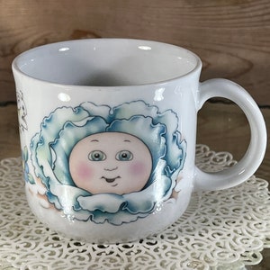 May include: White ceramic mug featuring a blue and white illustration of a baby doll's face within a flower. The mug has a handle.