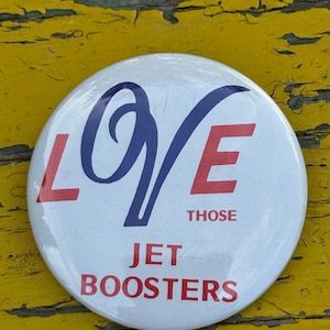 以下が含まれることがあります： 「Love Those Jet Boosters」と赤と青の文字が書かれた白いボタン。