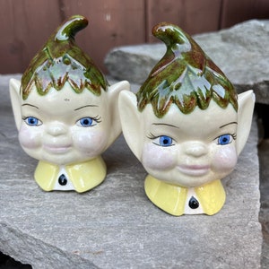 Puede incluir: Un par de saleros y pimenteros de cerámica en forma de elfo. Los saleros son blancos con sombreros verdes y cuellos amarillos. Tienen ojos azules y mejillas sonrosadas.