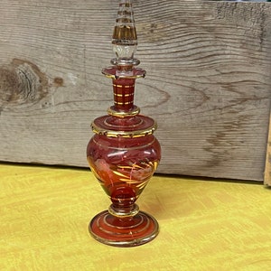 May include: A red glass bottle with gold accents and a clear glass stopper. The bottle has a round body and a narrow neck. The bottle is decorated with gold swirls and lines.