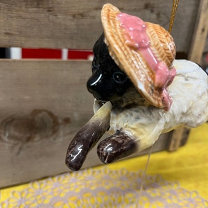 Puede incluir: Un adorno de cerámica de un gato negro con un sombrero de paja con una cinta rosa. El gato está colgado de una cadena dorada.