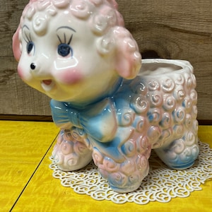 May include: A ceramic planter shaped like a pink and blue lamb with a blue bow. The lamb has a white face with pink cheeks and blue eyes.