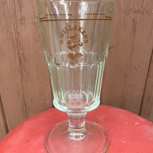 May include: A clear glass with a fluted design and a stem. The glass has a brown logo that says "G. Willakers" with a cartoon man wearing a hat and a moustache.