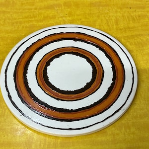 May include: A white ceramic coaster with concentric circles in brown, orange, and black.