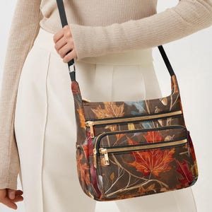 May include: A brown crossbody bag featuring a vibrant autumn leaf design. The bag has three compartments with gold zips and a black strap. The bag is approximately 25 cm wide and 20 cm tall.