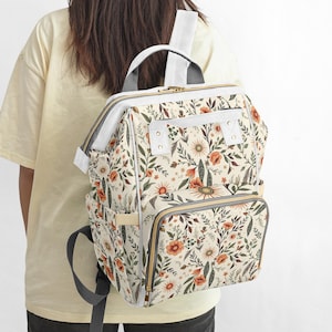 May include: A floral print backpack with a cream background, featuring orange and green flowers and foliage. The backpack has a top handle, adjustable straps, and multiple pockets. The design includes a front zippered compartment and a gray trim.