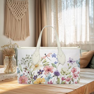 May include: White tote bag with a floral watercolor design featuring pink, purple, yellow, and blue flowers, a dragonfly, and a butterfly. The bag has cream-colored handles and a zipper closure. The bag is sitting on a wooden table.