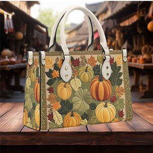 May include: A rectangular handbag with a fall theme. The bag features a pattern of pumpkins and autumn leaves in shades of orange, yellow, and green. It has white handles and trim, and is set on a wooden surface.