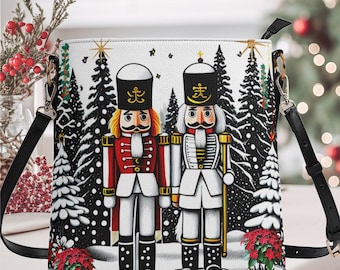 May include: White crossbody bag featuring a winter scene with two nutcracker figures. The bag has a black strap and silver-tone hardware. The nutcrackers are in red and white uniforms, standing in front of snow-covered trees. Red poinsettias are at the bottom.