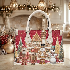 Peut inclure: Sac à main rouge et blanc sur le thème de Noël, avec une scène de village d'hiver représentant des bonhommes de neige, des casse-noisettes et un château. Le sac a une poignée et des accents blancs, avec une quincaillerie dorée.