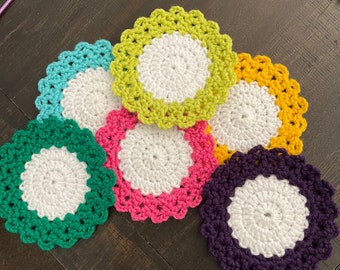 Op de afbeelding: Zes gehaakte onderzetters in verschillende kleuren, waaronder groen, paars, roze, geel en blauw. Elke onderzetter heeft een witte middelste cirkel.