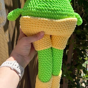 Op de afbeelding: Een handgemaakte gehaakte kikkerknuffel in heldergroen en geel. De kikker heeft een getextureerde uitstraling, met een ronde kop, armen, benen en een gele broek. Het speelgoed is zacht en knuffelbaar.