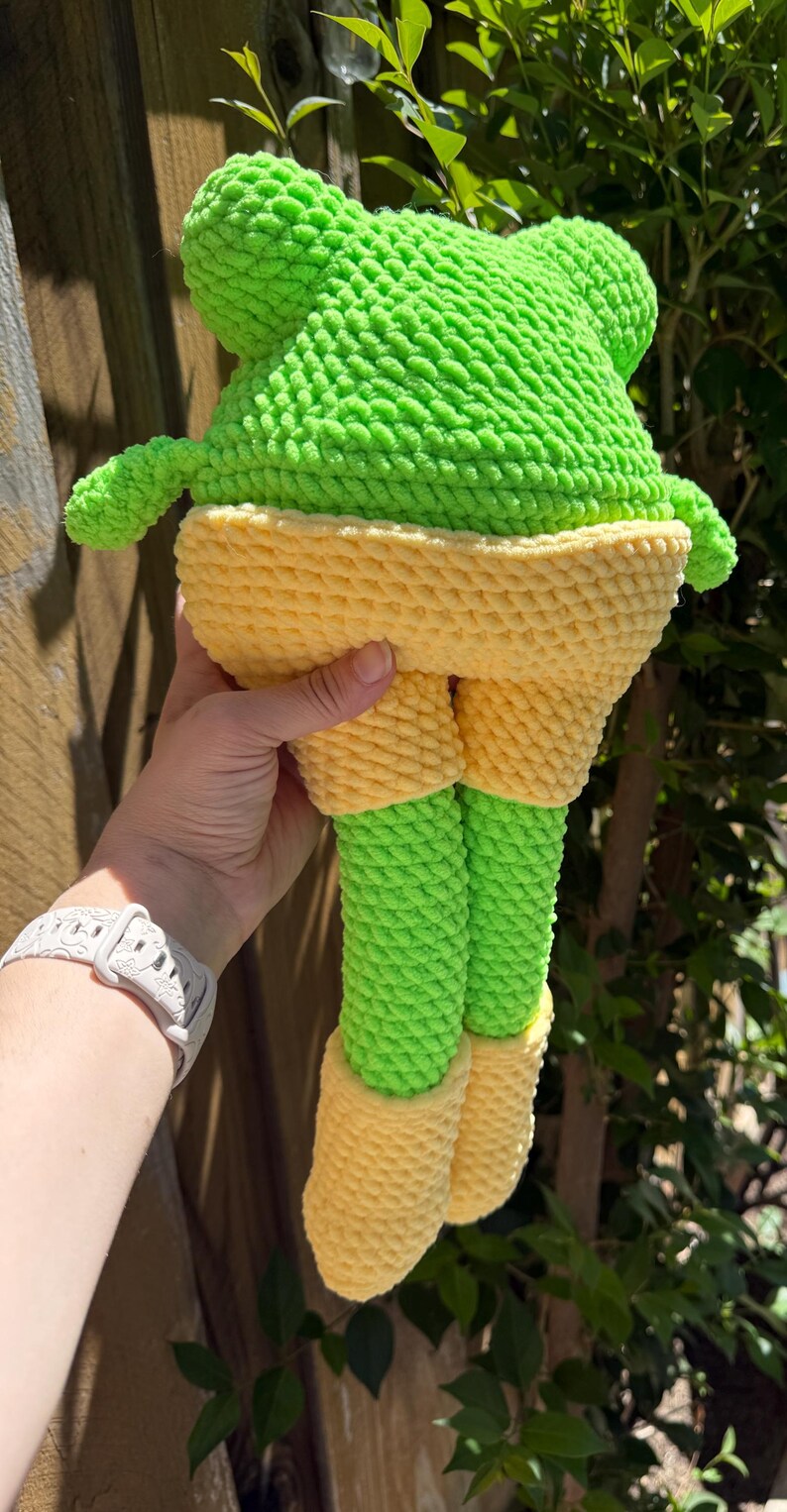 Op de afbeelding: Een handgemaakte gehaakte kikkerknuffel in heldergroen en geel. De kikker heeft een getextureerde uitstraling, met een ronde kop, armen, benen en een gele broek. Het speelgoed is zacht en knuffelbaar.