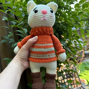 Pu&ograve; includere: Bambola a forma di gatto all'uncinetto fatta a mano con testa bianca, naso rosa e occhi neri. Il gatto indossa un maglione arancione con strisce verdi, pantaloncini bianchi e stivali marroni. La bambola &egrave; tenuta contro uno sfondo di fogliame verde.