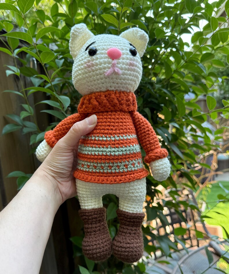 Pu&ograve; includere: Bambola a forma di gatto all'uncinetto fatta a mano con testa bianca, naso rosa e occhi neri. Il gatto indossa un maglione arancione con strisce verdi, pantaloncini bianchi e stivali marroni. La bambola &egrave; tenuta contro uno sfondo di fogliame verde.