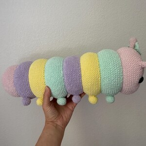 以下が含まれることがあります： パステルカラーの、かぎ針編みのイモムシのぬいぐるみ。イモムシはピンク、紫、黄色、ミントグリーンのセグメントが特徴です。小さな丸い足と、小さな触角と黒い目を持つ頭があります。おもちゃは手で持たれています。