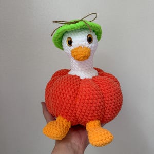 May include: A handmade crochet goose doll with a white head, orange beak and feet, and a bright green hat. The goose is wearing an orange body, giving it a pumpkin-like appearance. The doll is soft and plush.