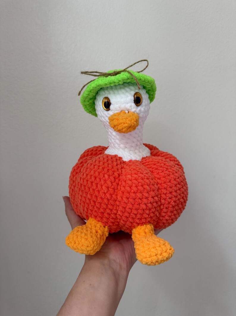 May include: A handmade crochet goose doll with a white head, orange beak and feet, and a bright green hat. The goose is wearing an orange body, giving it a pumpkin-like appearance. The doll is soft and plush.
