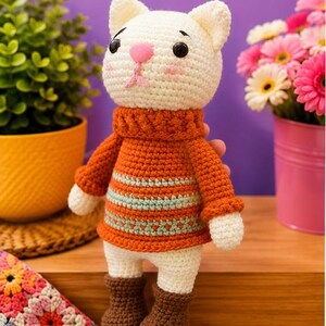 Pu&ograve; includere: Bambola a forma di gatto all'uncinetto fatta a mano con testa bianca, naso rosa e occhi neri. Indossa un maglione arancione con strisce blu e bianche e stivali marroni. La bambola &egrave; su una superficie di legno.