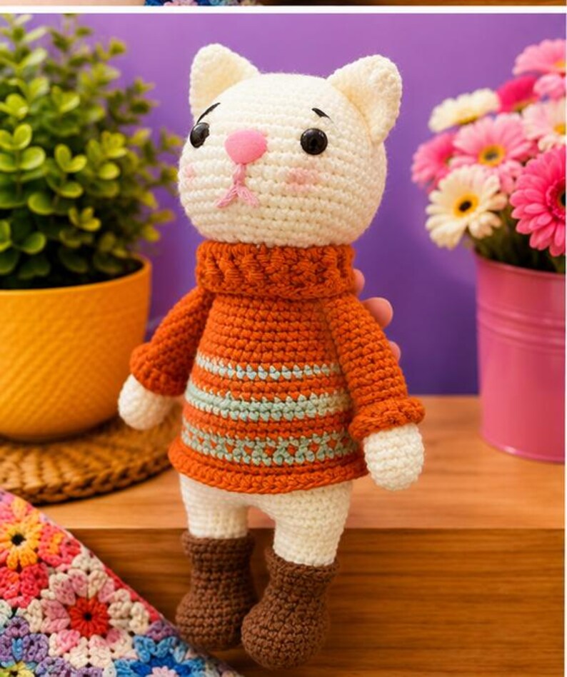 Pu&ograve; includere: Bambola a forma di gatto all'uncinetto fatta a mano con testa bianca, naso rosa e occhi neri. Indossa un maglione arancione con strisce blu e bianche e stivali marroni. La bambola &egrave; su una superficie di legno.