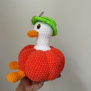 May include: A handmade crochet duck with a white head, orange beak and feet, and a bright orange body. The duck wears a green hat. The duck is a whimsical, soft toy.