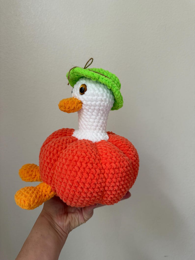 May include: A handmade crochet duck with a white head, orange beak and feet, and a bright orange body. The duck wears a green hat. The duck is a whimsical, soft toy.