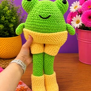 Op de afbeelding: Een handgemaakte, groene gehaakte kikkerpop met gele shorts en laarzen. De kikker heeft zwarte knoopogen en een lachende mond. De pop wordt vastgehouden tegen een paarse achtergrond, met planten en bloemen op de achtergrond.