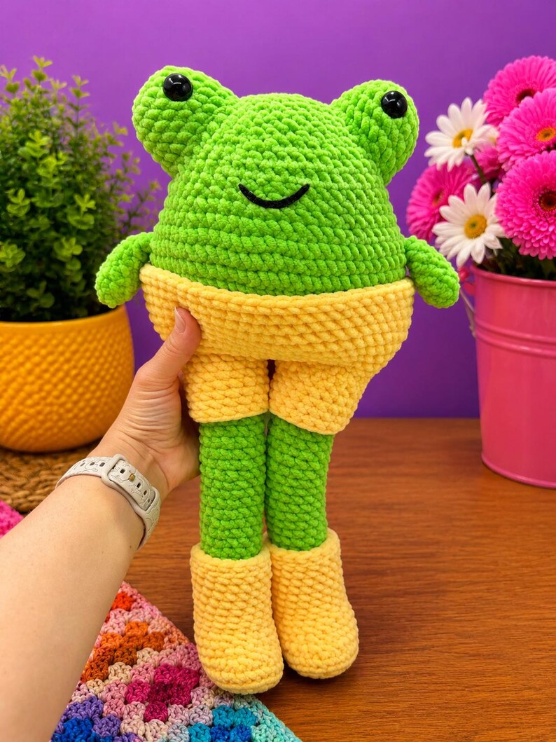Op de afbeelding: Een handgemaakte, groene gehaakte kikkerpop met gele shorts en laarzen. De kikker heeft zwarte knoopogen en een lachende mond. De pop wordt vastgehouden tegen een paarse achtergrond, met planten en bloemen op de achtergrond.