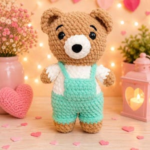 K&ouml;nnte beinhalten: Ein geh&auml;kelter Teddyb&auml;r mit hellbraunem Fell, wei&szlig;er Schnauze und schwarzen Kunststoffaugen. Der B&auml;r tr&auml;gt wei&szlig;e kurze &Auml;rmel und eine mintgr&uuml;ne Latzhose mit quadratischem Latz. Das Pl&uuml;schtier besteht aus weichem, strukturiertem Garn.