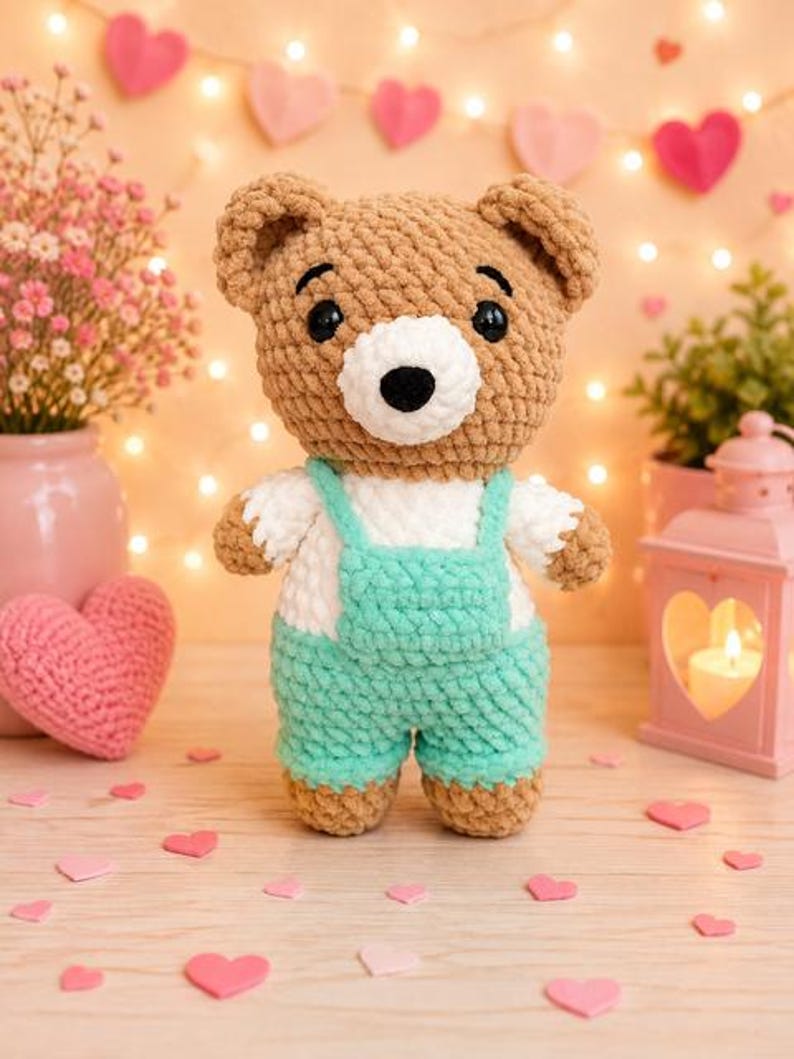 K&ouml;nnte beinhalten: Ein geh&auml;kelter Teddyb&auml;r mit hellbraunem Fell, wei&szlig;er Schnauze und schwarzen Kunststoffaugen. Der B&auml;r tr&auml;gt wei&szlig;e kurze &Auml;rmel und eine mintgr&uuml;ne Latzhose mit quadratischem Latz. Das Pl&uuml;schtier besteht aus weichem, strukturiertem Garn.