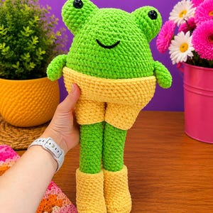 Op de afbeelding: Een handgemaakte, groene gehaakte kikkerpop met gele shorts en laarzen. De kikker heeft zwarte ogen en een lachende mond. De pop wordt vastgehouden tegen een paarse achtergrond met planten en bloemen.