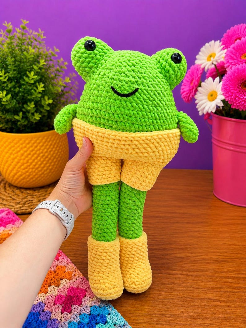 Op de afbeelding: Een handgemaakte, groene gehaakte kikkerpop met gele shorts en laarzen. De kikker heeft zwarte ogen en een lachende mond. De pop wordt vastgehouden tegen een paarse achtergrond met planten en bloemen.