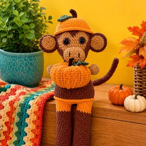 May include: A brown and orange crocheted monkey toy wearing a matching orange hat with a small green leaf. The monkey has large dark eyes and is holding a textured orange crocheted pumpkin with a green stem and leaf in its hands.