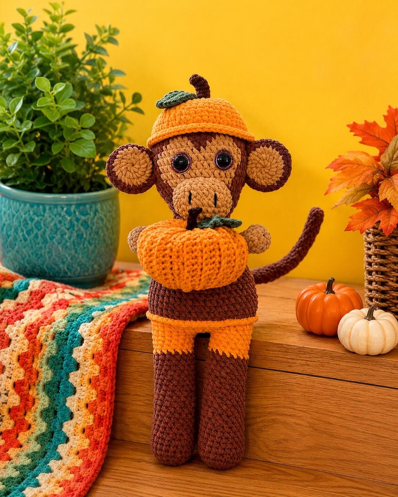 May include: A brown and orange crocheted monkey toy wearing a matching orange hat with a small green leaf. The monkey has large dark eyes and is holding a textured orange crocheted pumpkin with a green stem and leaf in its hands.