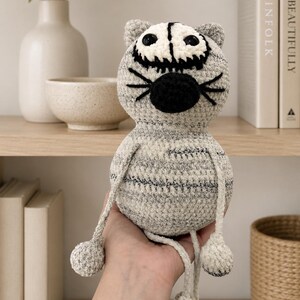 May include: A gray and white crocheted cat plush toy with a round head, black embroidered whiskers, a black nose, and large dark eyes. The body features a mottled gray yarn texture with thin horizontal stripes, long dangling arms, and legs.