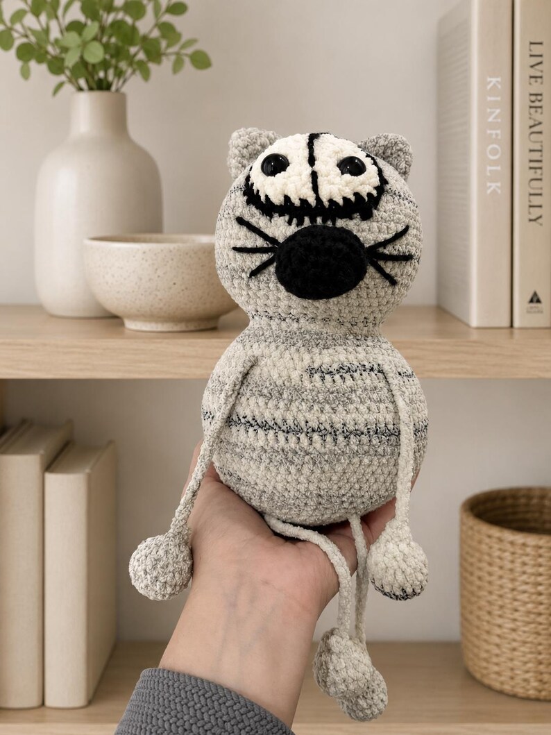 May include: A gray and white crocheted cat plush toy with a round head, black embroidered whiskers, a black nose, and large dark eyes. The body features a mottled gray yarn texture with thin horizontal stripes, long dangling arms, and legs.