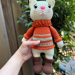 Pu&ograve; includere: Bambola a forma di gatto all'uncinetto fatta a mano con testa bianca, naso rosa e occhi neri. Il gatto indossa un maglione arancione con strisce verdi e bianche e stivali marroni. La bambola &egrave; alta circa 20 cm.