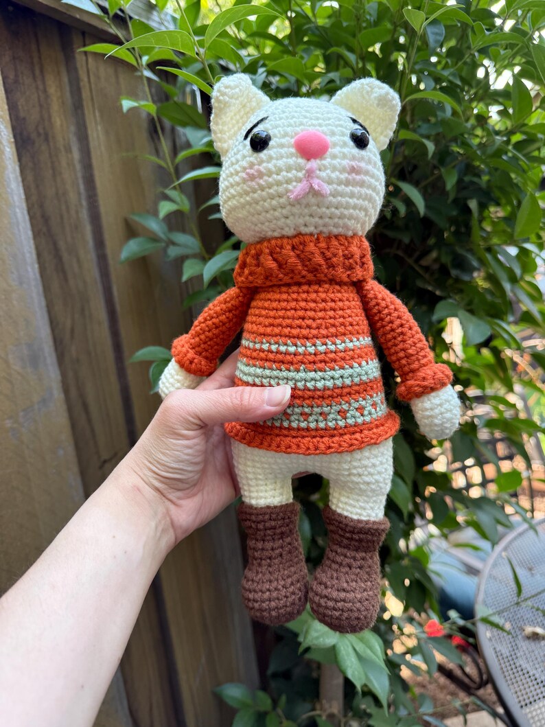 Pu&ograve; includere: Bambola a forma di gatto all'uncinetto fatta a mano con testa bianca, naso rosa e occhi neri. Il gatto indossa un maglione arancione con strisce verdi e bianche e stivali marroni. La bambola &egrave; alta circa 20 cm.