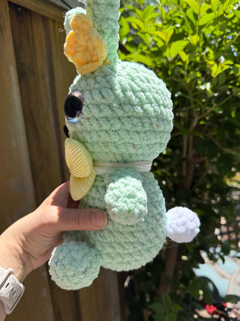 Pode incluir: Um coelho de croch&ecirc; verde claro feito &agrave; m&atilde;o, com uma coroa e la&ccedil;o amarelos. O coelho tem olhos grandes e reflexivos e uma pequena cauda de pompom branca. O fio macio e texturizado tem manchas de cor, dando-lhe uma apar&ecirc;ncia caprichosa.