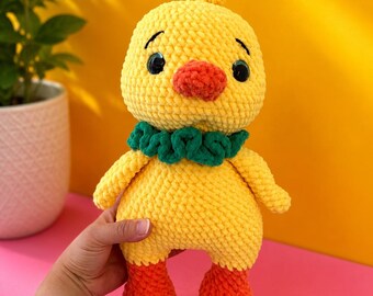 Può includere: Un pulcino all'uncinetto giallo fatto a mano con zampe arancioni e un becco rosso. Il pulcino ha un colletto verde e occhi neri. Il pulcino è tenuto davanti a uno sfondo giallo e rosa.