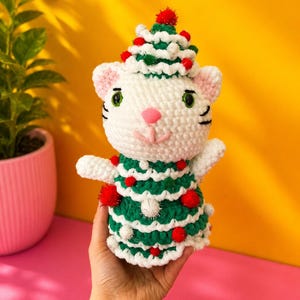 Pode incluir: Um gato de croch&ecirc; branco feito &agrave; m&atilde;o usando uma fantasia de &aacute;rvore de Natal verde e branca com pompons vermelhos. O gato tem olhos verdes, nariz rosa e bigodes pretos. O gato est&aacute; sendo segurado na m&atilde;o de uma pessoa contra um fundo amarelo e rosa.