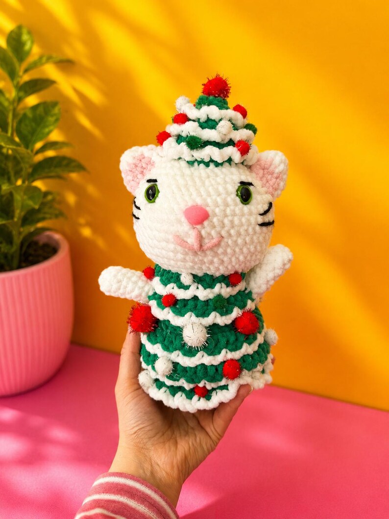 Pode incluir: Um gato de croch&ecirc; branco feito &agrave; m&atilde;o usando uma fantasia de &aacute;rvore de Natal verde e branca com pompons vermelhos. O gato tem olhos verdes, nariz rosa e bigodes pretos. O gato est&aacute; sendo segurado na m&atilde;o de uma pessoa contra um fundo amarelo e rosa.